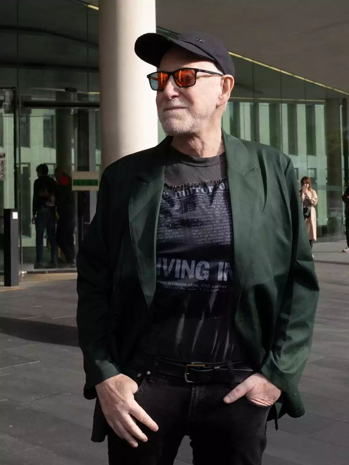 Un hombre con gafas de sol y gorra negra, vistiendo una camiseta negra con texto y una chaqueta verde, está de pie frente a un edificio con paredes de vidrio.