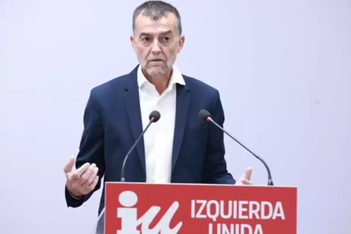 A man standing behind a podium with the Izquierda Unida logo, speaking in front of two microphones.