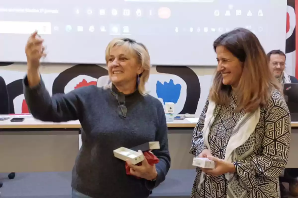 Dos mujeres sonríen mientras una de ellas sostiene una caja y parece estar tomando una foto con un teléfono móvil en un entorno de oficina o conferencia.