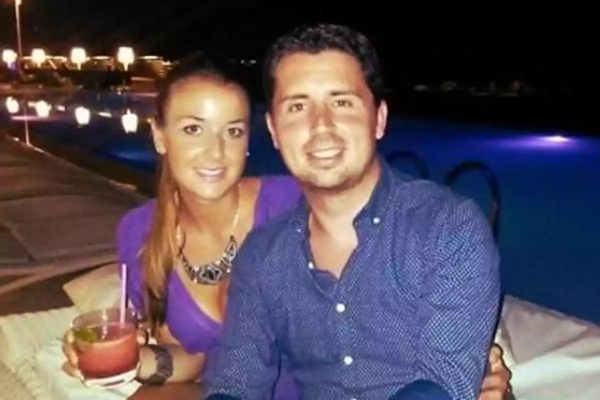 Una pareja sonriente sentada junto a una piscina iluminada por la noche, la mujer sostiene una bebida con pajilla.