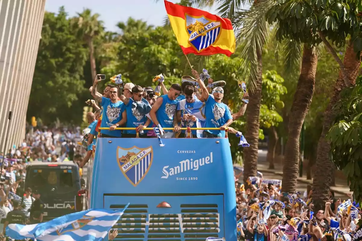 Un grupo de personas celebra en la parte superior de un autobús descapotable con banderas y camisetas del Málaga C.F. mientras una multitud los acompaña en la calle.