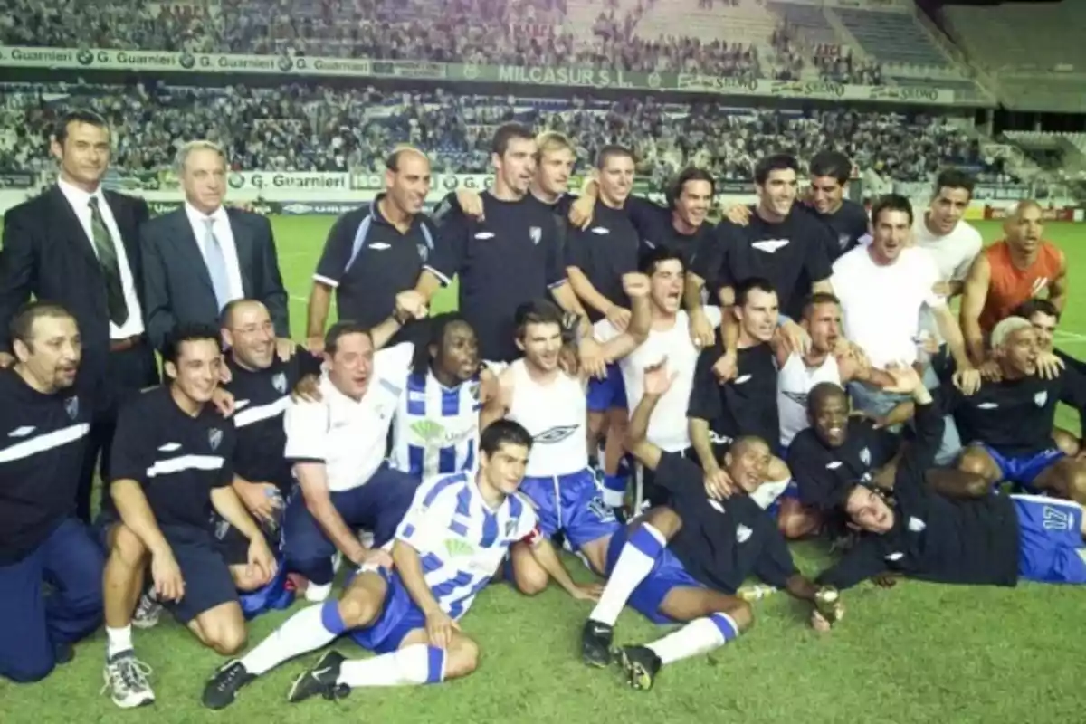 Un grupo de jugadores de fútbol y personal técnico posan juntos en un campo de fútbol, celebrando un logro deportivo frente a una multitud en las gradas.