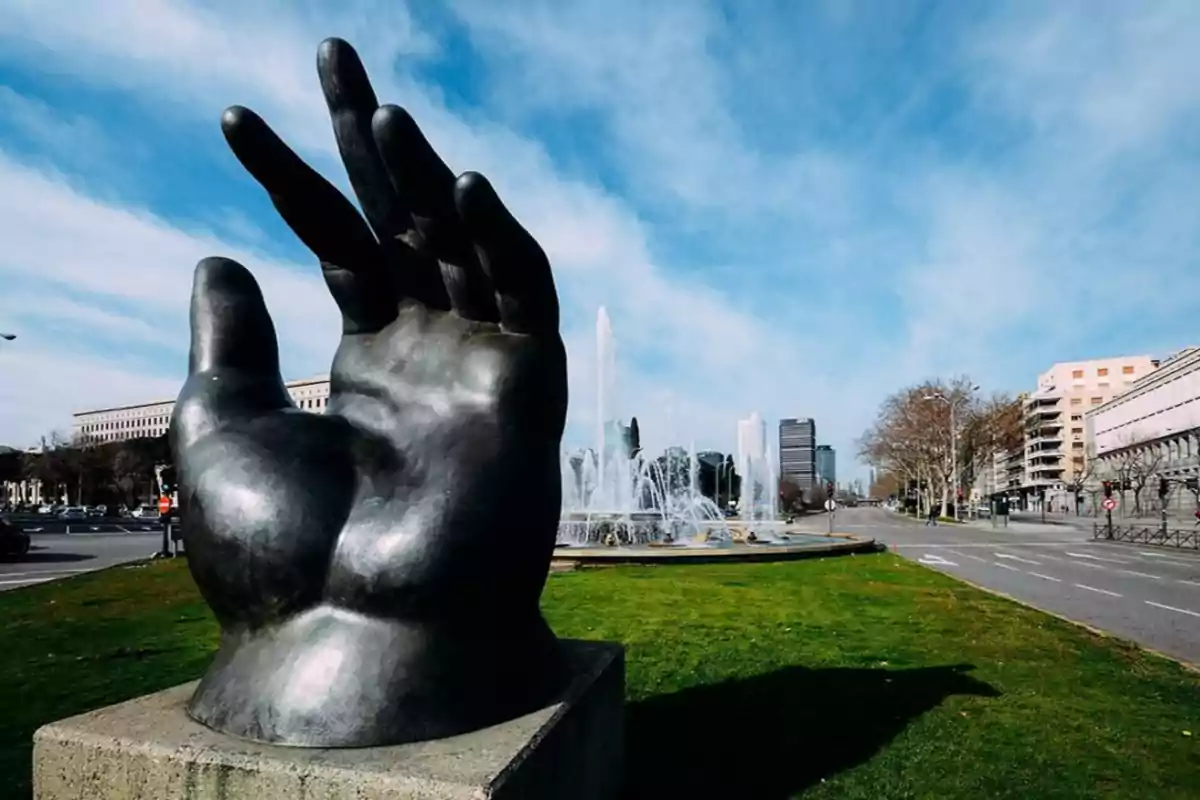 Escultura de una mano gigante en un parque urbano con una fuente y edificios al fondo.