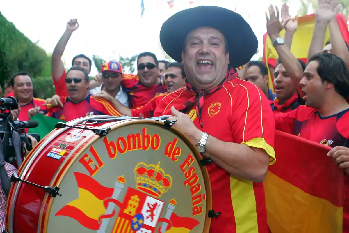 Un grupo de aficionados con camisetas rojas y amarillas celebra con entusiasmo mientras uno de ellos toca un bombo decorado con la bandera de España.