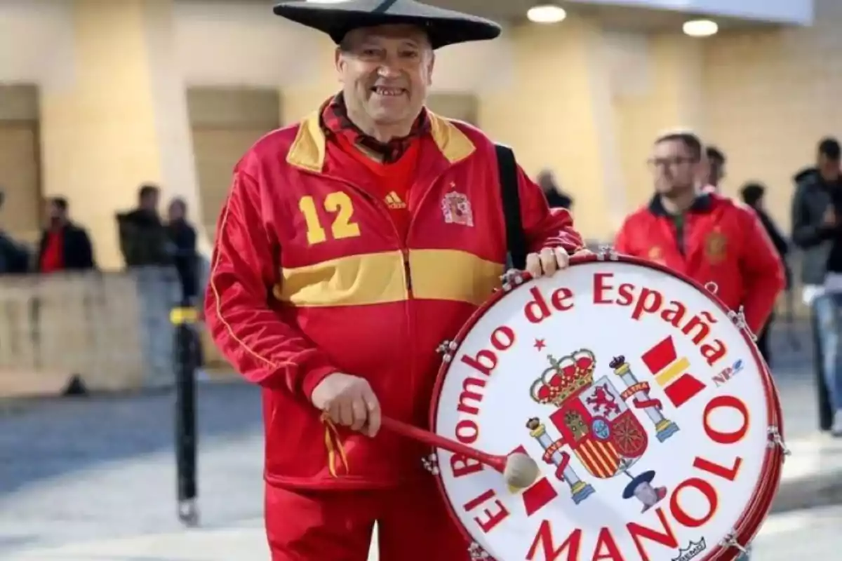 Un hombre con un sombrero y un chándal rojo y amarillo sostiene un gran bombo decorado con símbolos de España. Un hombre con un sombrero y un chándal rojo y amarillo sostiene un gran bombo decorado con símbolos de España.