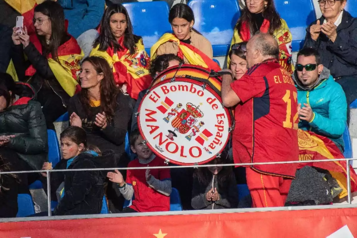 Un grupo de personas en un estadio, algunas con banderas de España, observan a un hombre tocando un bombo que tiene escrito Un grupo de personas en un estadio, algunas con banderas de España, observan a un hombre tocando un bombo que tiene escrito