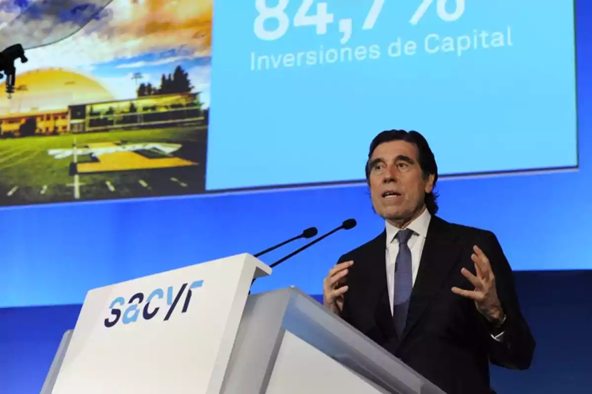 Un hombre en traje y corbata está dando una presentación en un podio con el logotipo de Sacyr, mientras en la pantalla detrás de él se muestra una imagen de un campo deportivo y un texto que dice "84,7% Inversiones de Capital".