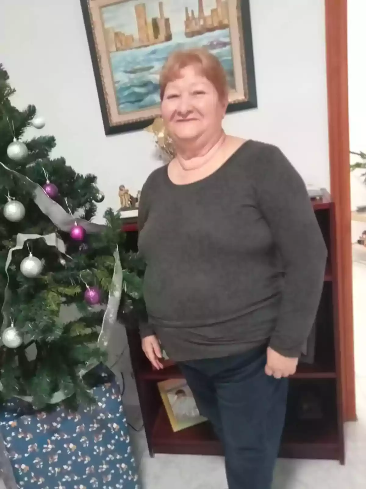 Una mujer de pie junto a un árbol de Navidad decorado con esferas plateadas y moradas, con una pintura de un paisaje urbano en la pared detrás de ella.