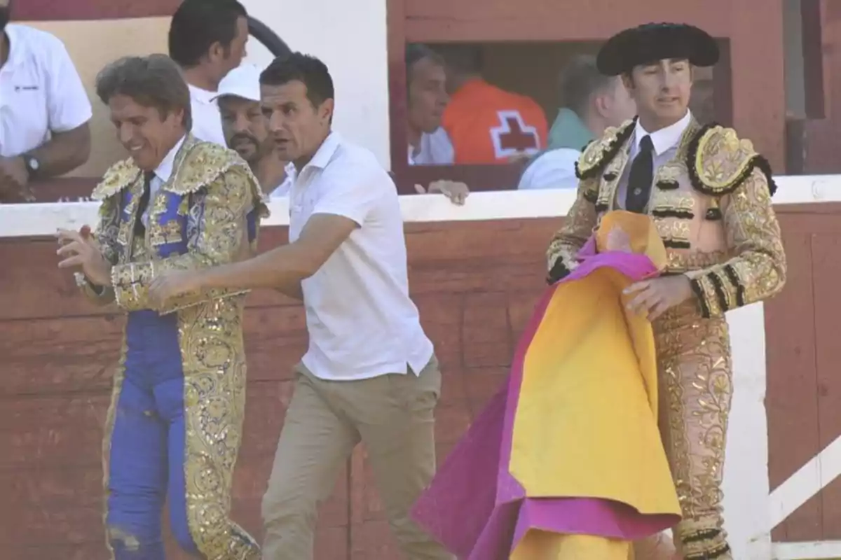 Dos toreros y un hombre con camiseta blanca están en una plaza de toros, uno de los toreros lleva un traje azul y dorado, mientras que el otro lleva un traje dorado y negro y sostiene un capote.