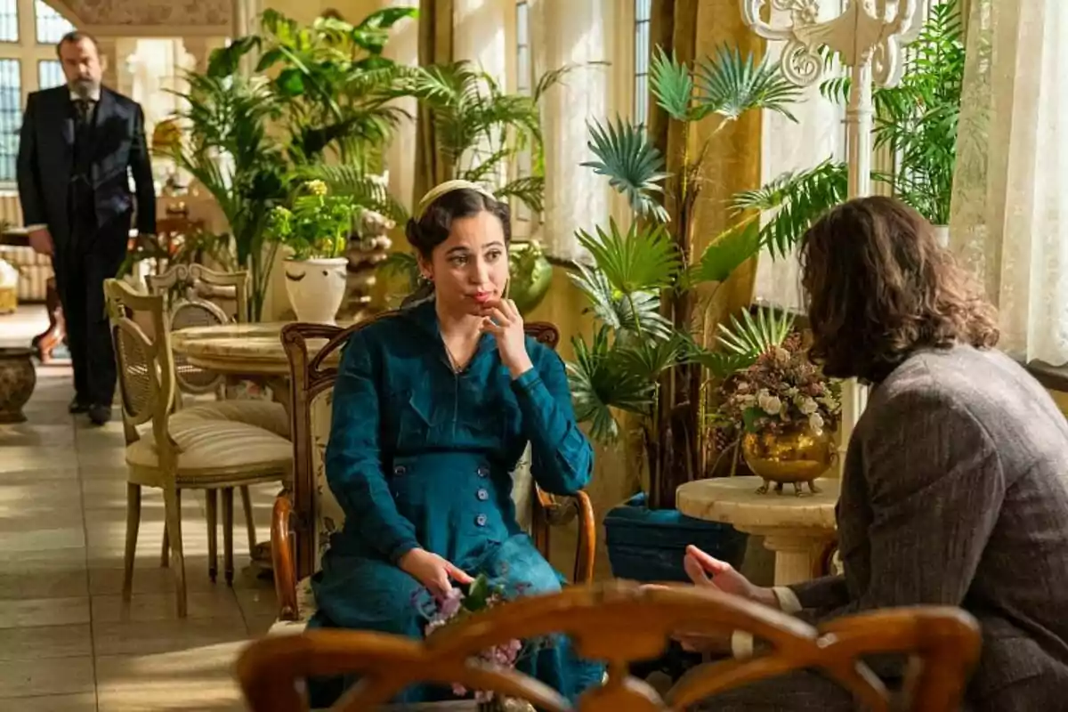 Carmen Asecas con vestido azul está sentada en una silla en una habitación decorada con plantas mientras conversa con Ibrahim Al Shami de cabello largo; al fondo, Manuel Regueiro observa la escena en La Promesa. Carmen Asecas con vestido azul está sentada en una silla en una habitación decorada con plantas mientras conversa con Ibrahim Al Shami de cabello largo; al fondo, Manuel Regueiro observa la escena en La Promesa.