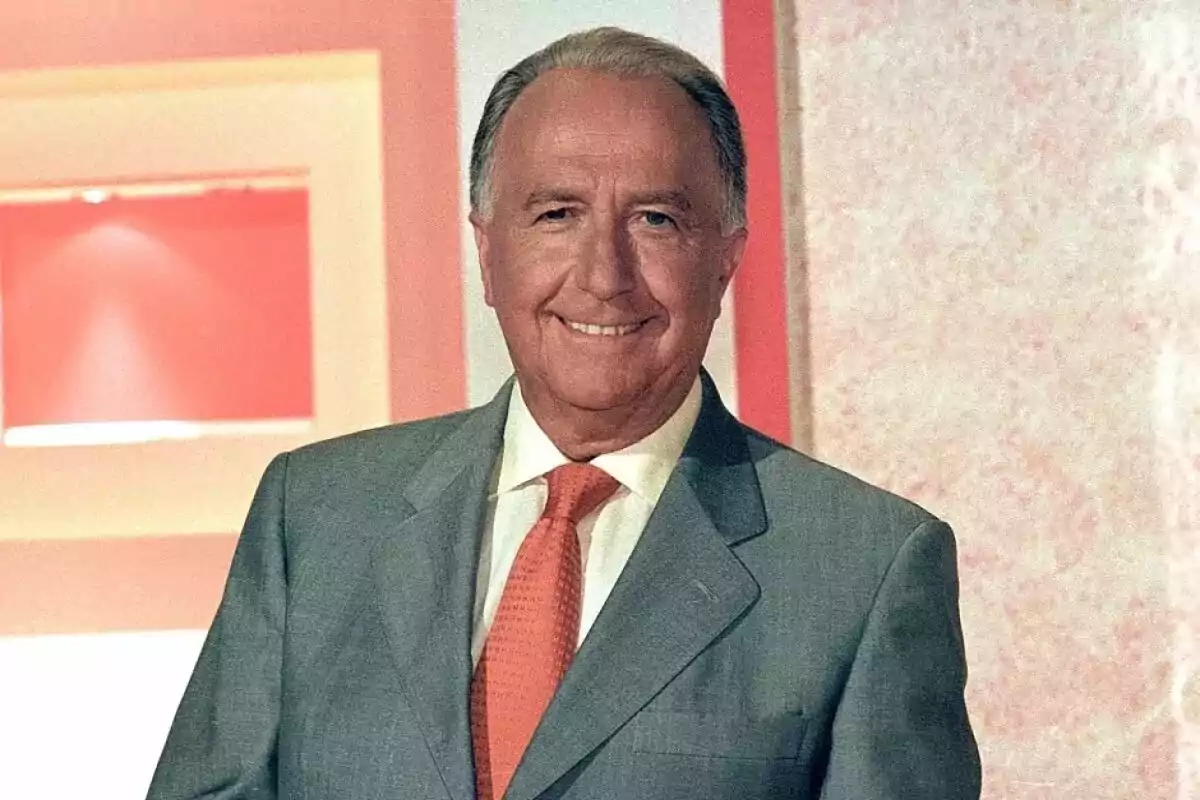 Un hombre mayor con traje gris y corbata naranja sonríe frente a un fondo rojo y blanco.