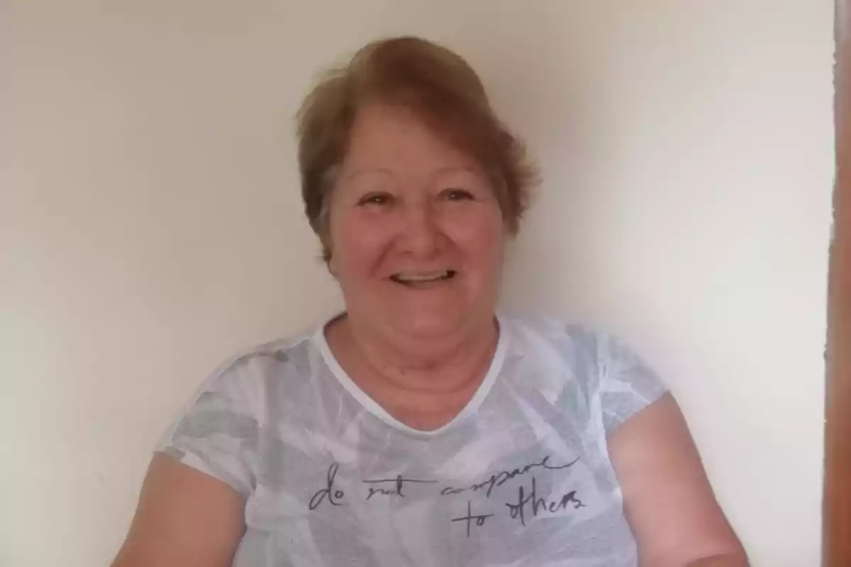 Una mujer mayor sonriente con cabello corto y una camiseta blanca con la frase "do not compare to others"