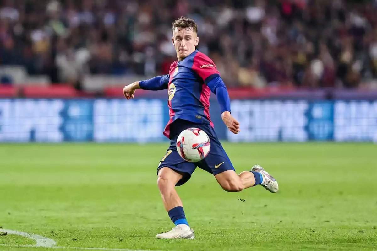 Jugador de fútbol en acción durante un partido con uniforme azul y rojo en un estadio.