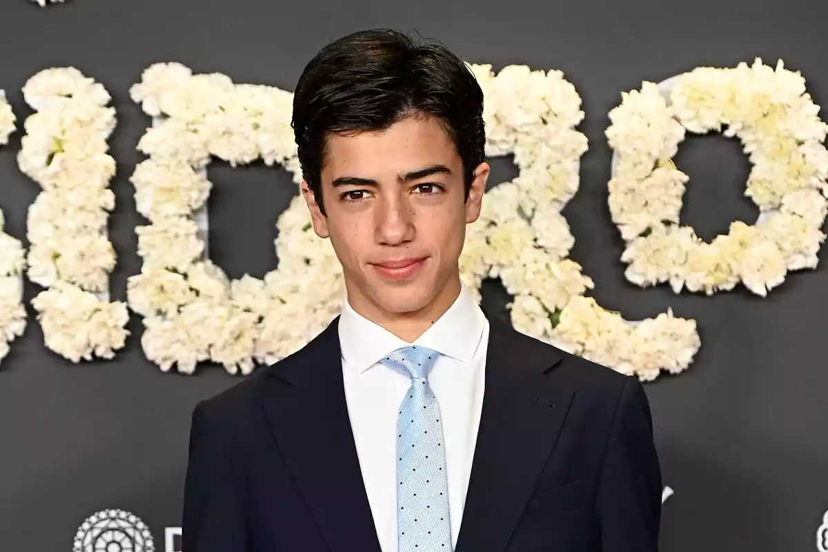 Un hombre joven con traje oscuro y corbata azul claro posando frente a un fondo decorado con flores blancas.