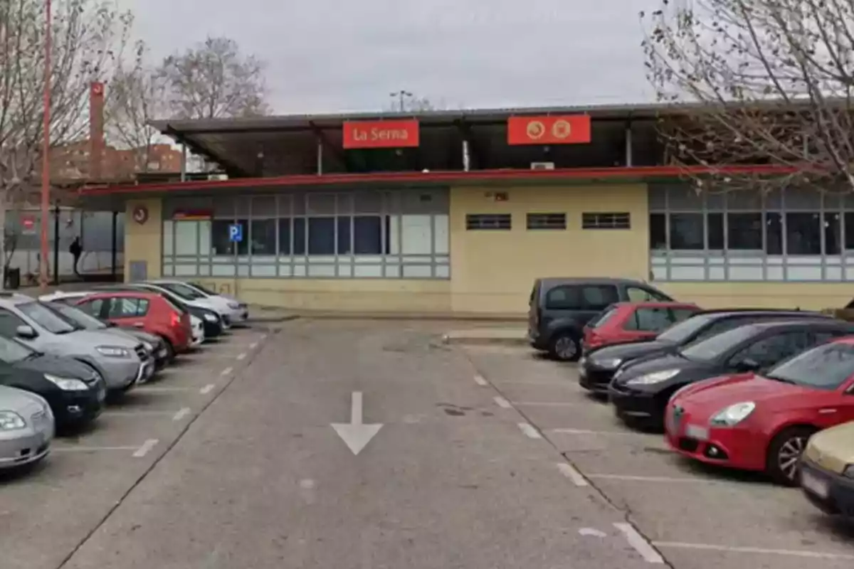 La imagen muestra un estacionamiento con varios automóviles aparcados frente a un edificio con un letrero que dice 