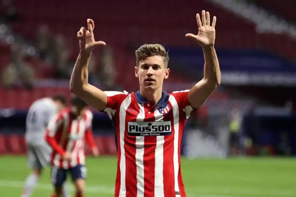 Jugador de fútbol con uniforme del Atlético de Madrid levantando las manos en señal de celebración en un estadio.