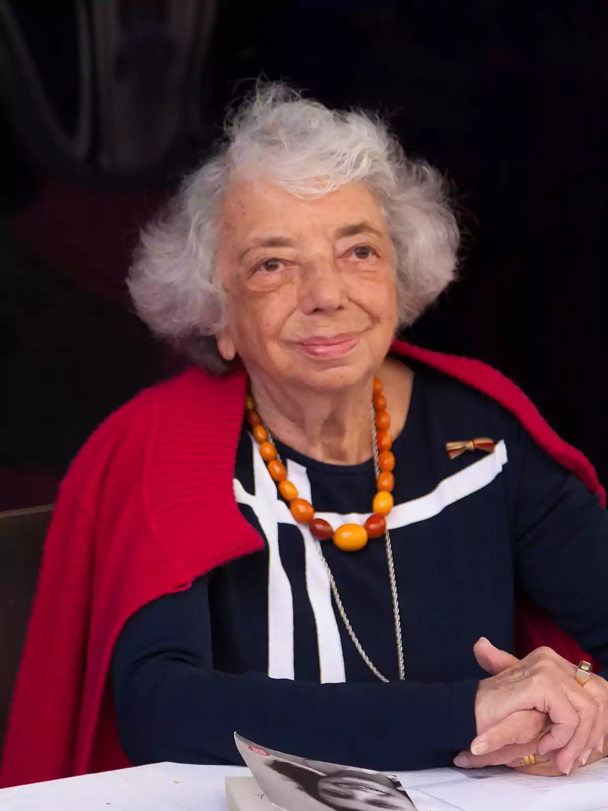 Una persona mayor con cabello canoso y un collar naranja, vestida con un suéter rojo y una blusa azul con detalles blancos, está sentada frente a una mesa con un libro. Una persona mayor con cabello canoso y un collar naranja, vestida con un suéter rojo y una blusa azul con detalles blancos, está sentada frente a una mesa con un libro.