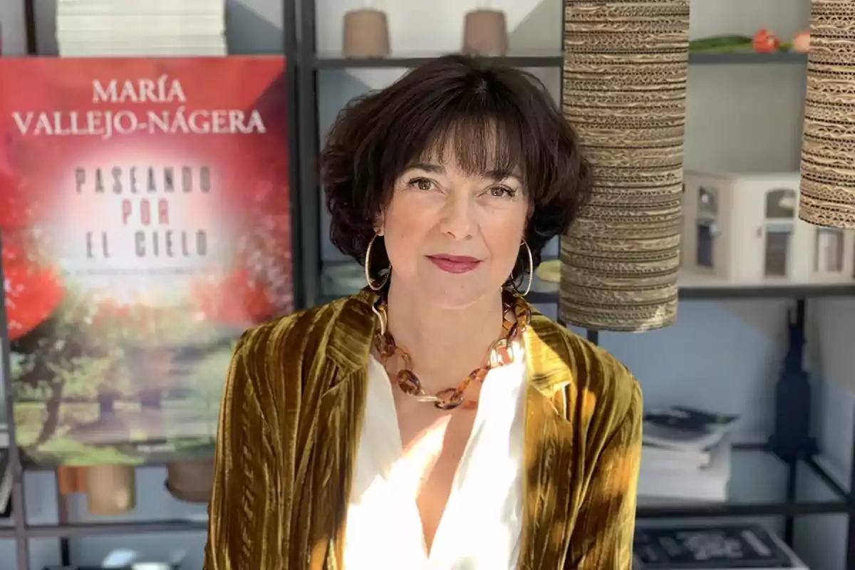 Una mujer de cabello oscuro y corto, con un collar y pendientes grandes, está de pie frente a un cartel que dice 