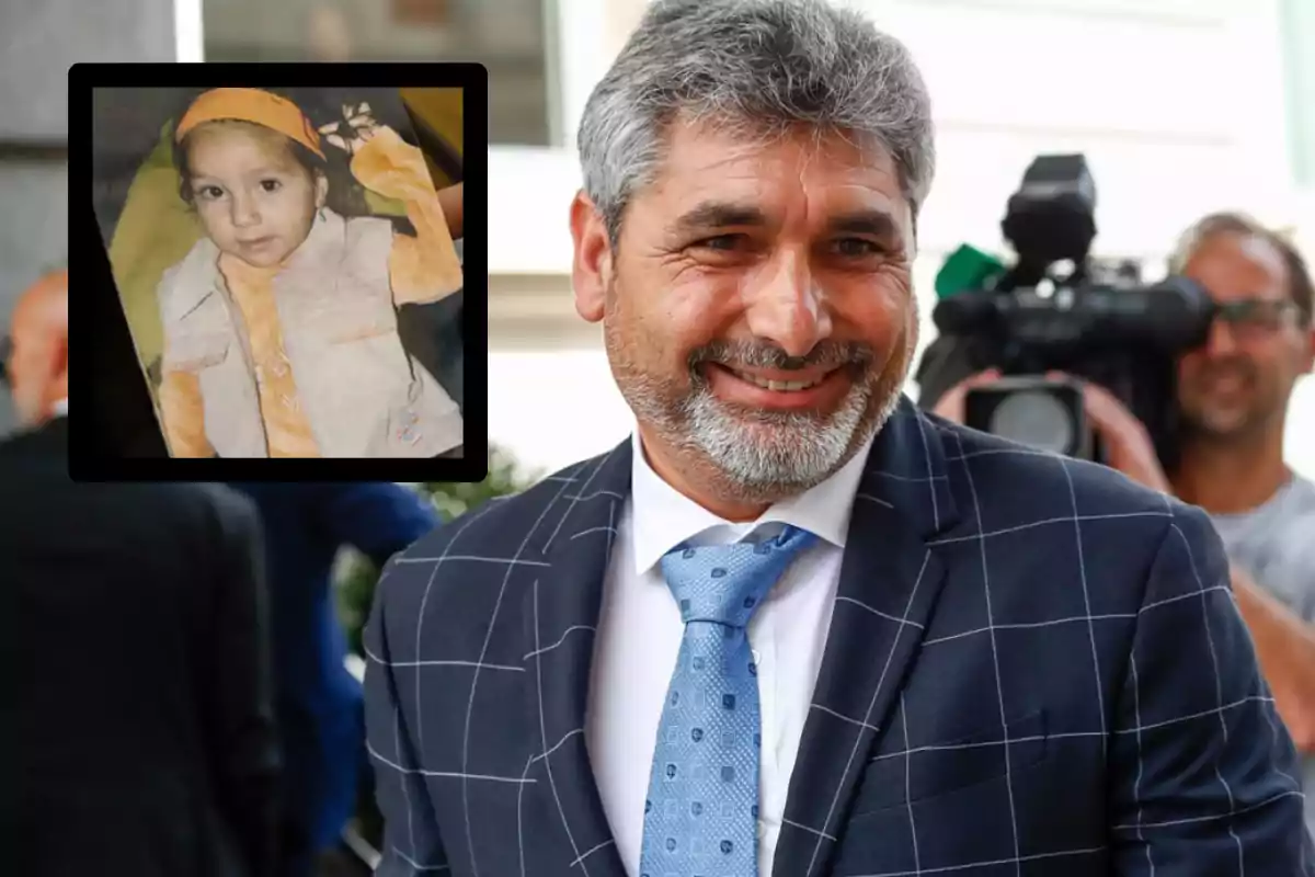 Un hombre sonriente con barba y traje a cuadros posa frente a una cámara, mientras sostiene una foto de una niña pequeña vestida de naranja y beige.