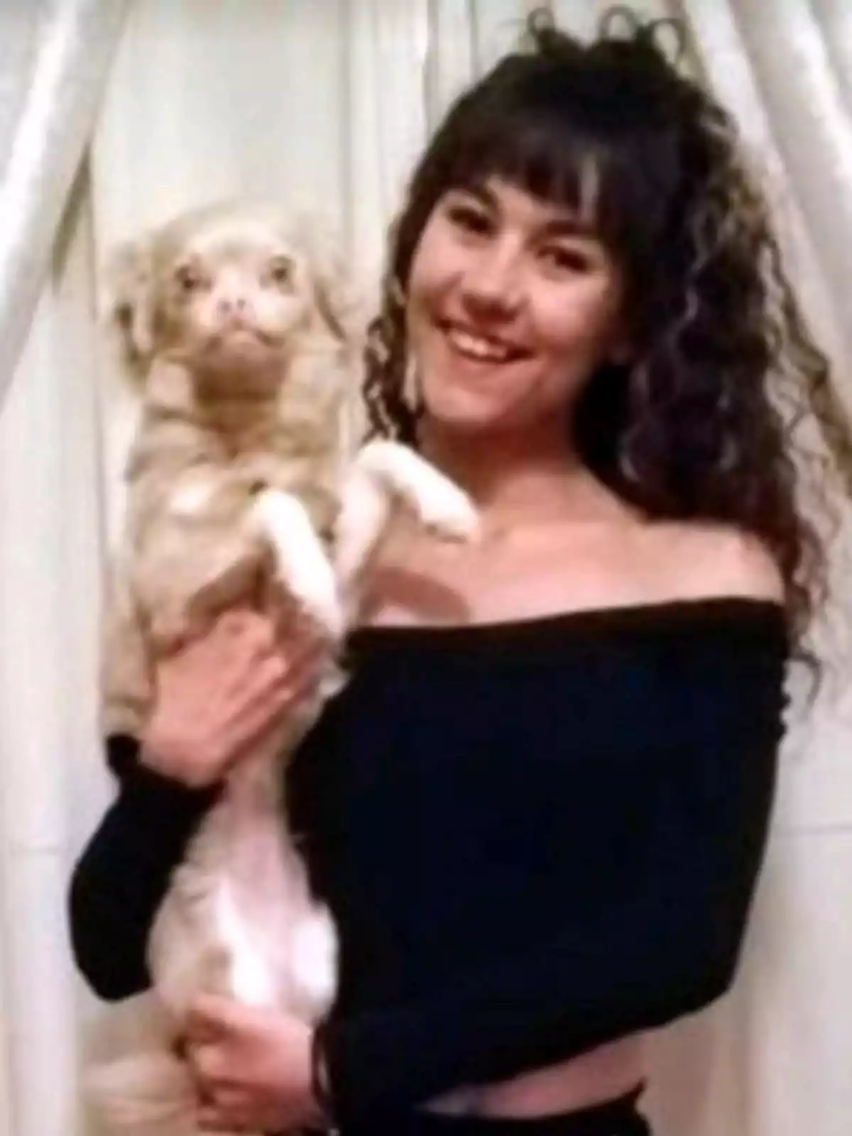 Una mujer sonriente con cabello rizado sostiene a un perro pequeño de color claro frente a una cortina blanca.