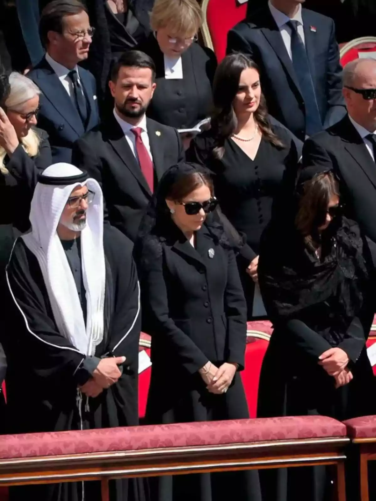 A group of people dressed in black, some wearing sunglasses, is standing at an outdoor formal event. A group of people dressed in black, some wearing sunglasses, is standing at an outdoor formal event.