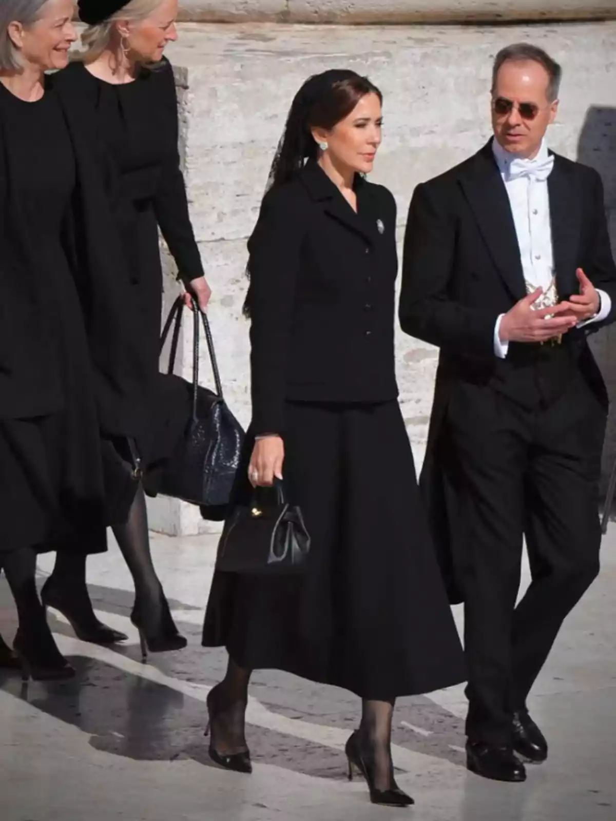 A group of people dressed in black walks on a stone surface, with a woman in a veil and black purse standing out next to a man in a tuxedo. A group of people dressed in black walks on a stone surface, with a woman in a veil and black purse standing out next to a man in a tuxedo.