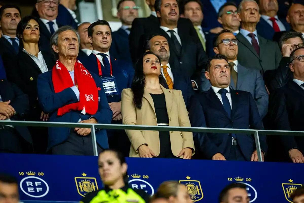 Un grupo de personas vestidas formalmente observa atentamente desde las gradas de un estadio, destacando un hombre con una bufanda roja. Un grupo de personas vestidas formalmente observa atentamente desde las gradas de un estadio, destacando un hombre con una bufanda roja.