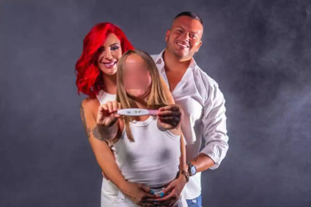 Tres personas posan juntas, una mujer con cabello rojo y un hombre abrazan a una mujer que sostiene una prueba de embarazo.