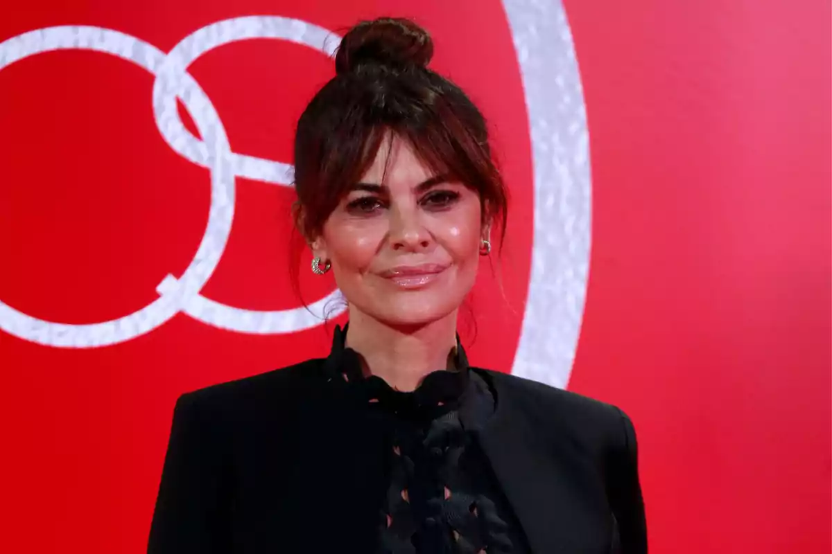 Una persona con cabello recogido y vestimenta oscura posando frente a un fondo rojo con un diseño circular.