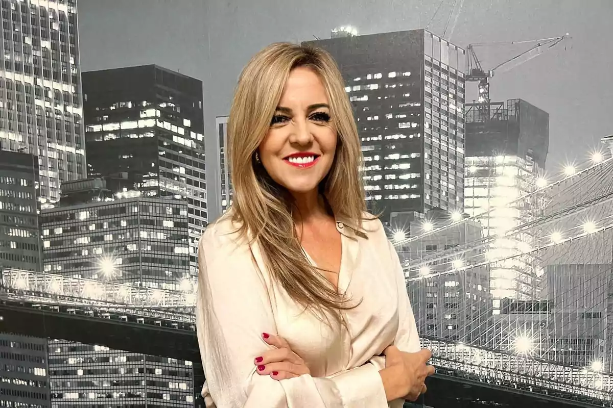 Una mujer sonriente con cabello rubio y camisa clara posando frente a un fondo de edificios iluminados.