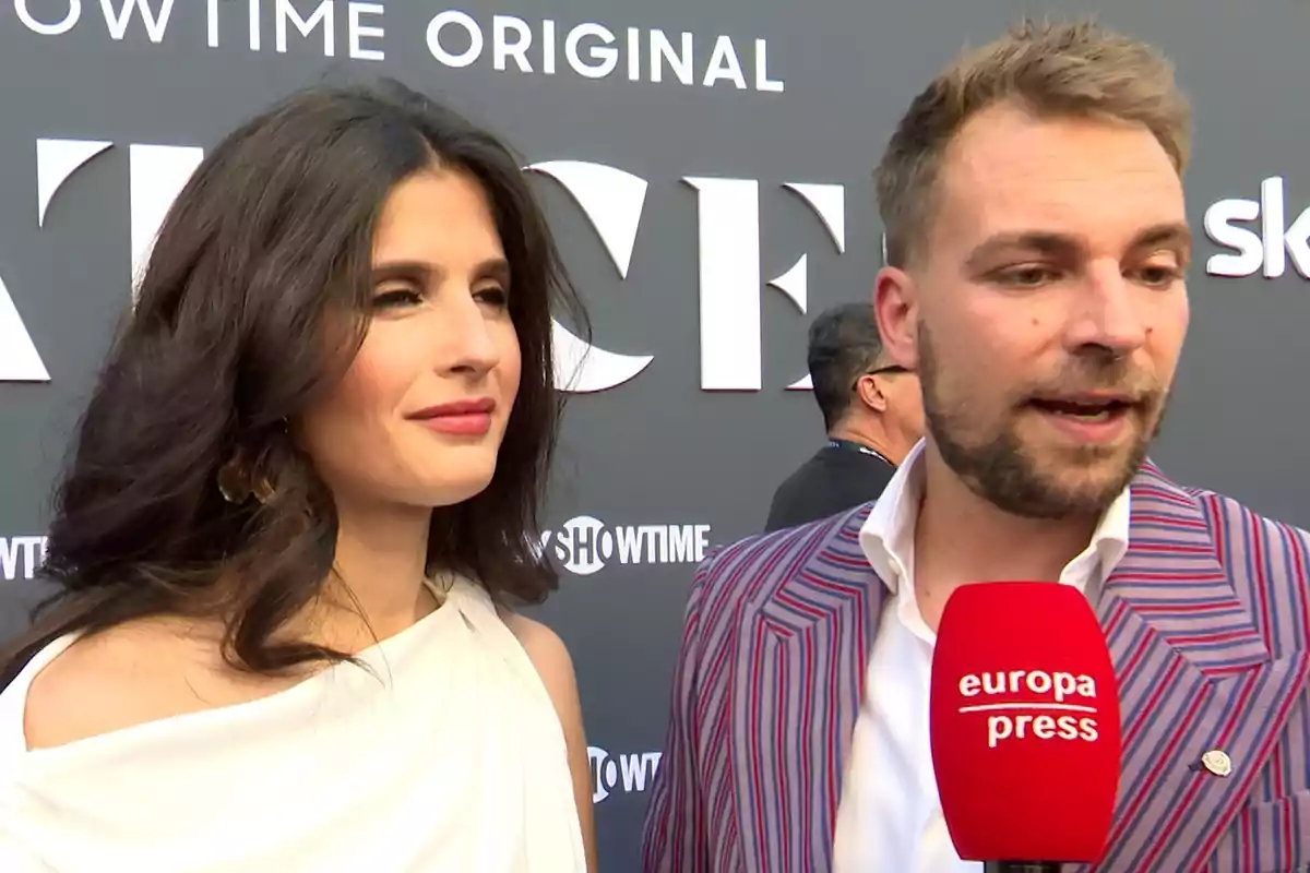 María Sánchez con vestido blanco y José María Almoguera con saco de rayas hablan frente a un micrófono rojo de europa press en un evento con fondo de letras grandes. María Sánchez con vestido blanco y José María Almoguera con saco de rayas hablan frente a un micrófono rojo de europa press en un evento con fondo de letras grandes.