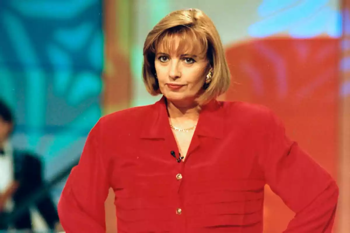Mujer con cabello corto y rubio, vestida con una blusa roja, en un set de televisión con fondo colorido.