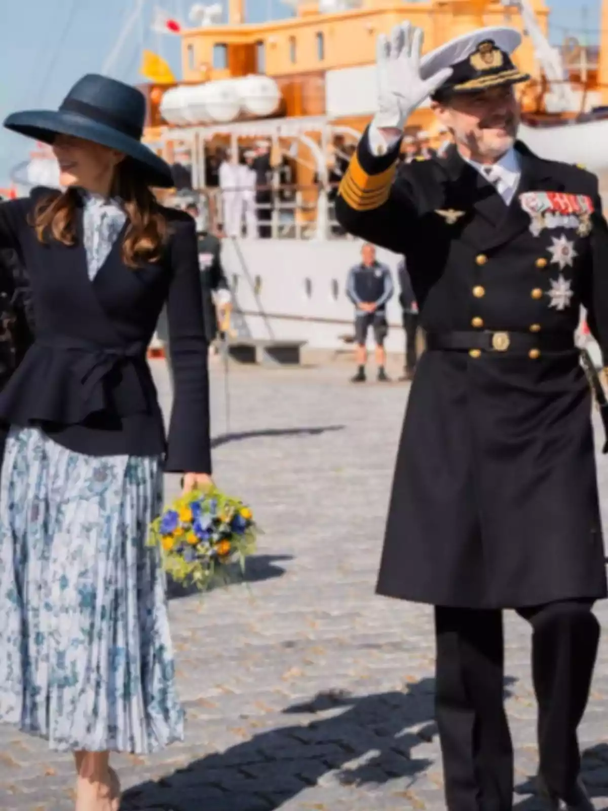 Una mujer con sombrero y vestido floral sostiene un ramo de flores mientras camina junto a un hombre con uniforme naval que saluda, con un barco de fondo. Una mujer con sombrero y vestido floral sostiene un ramo de flores mientras camina junto a un hombre con uniforme naval que saluda, con un barco de fondo.