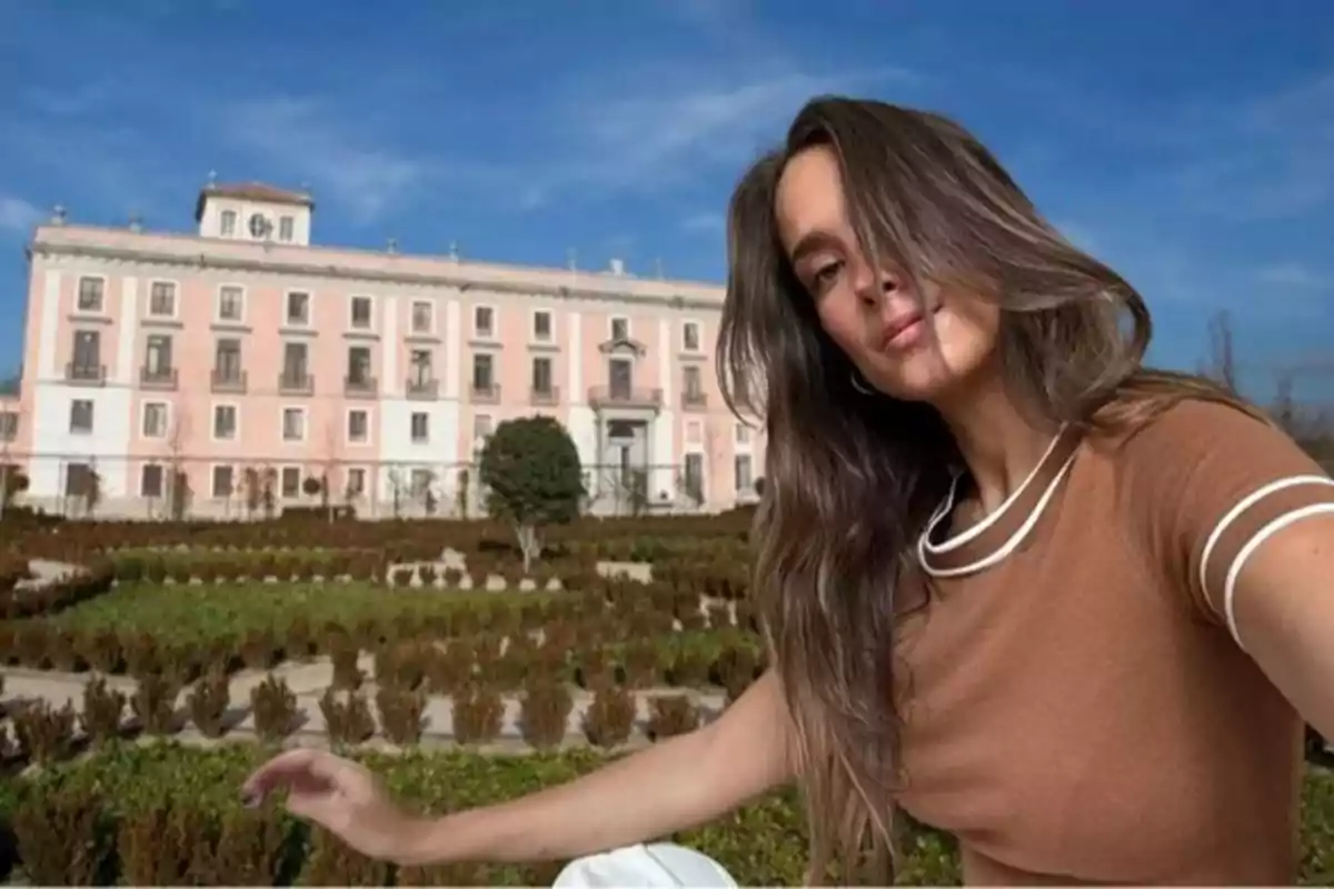 Una mujer con cabello largo y suelto se toma una selfie frente a un edificio grande y elegante con una fachada de color rosa y blanco, rodeado de jardines bien cuidados bajo un cielo azul claro.