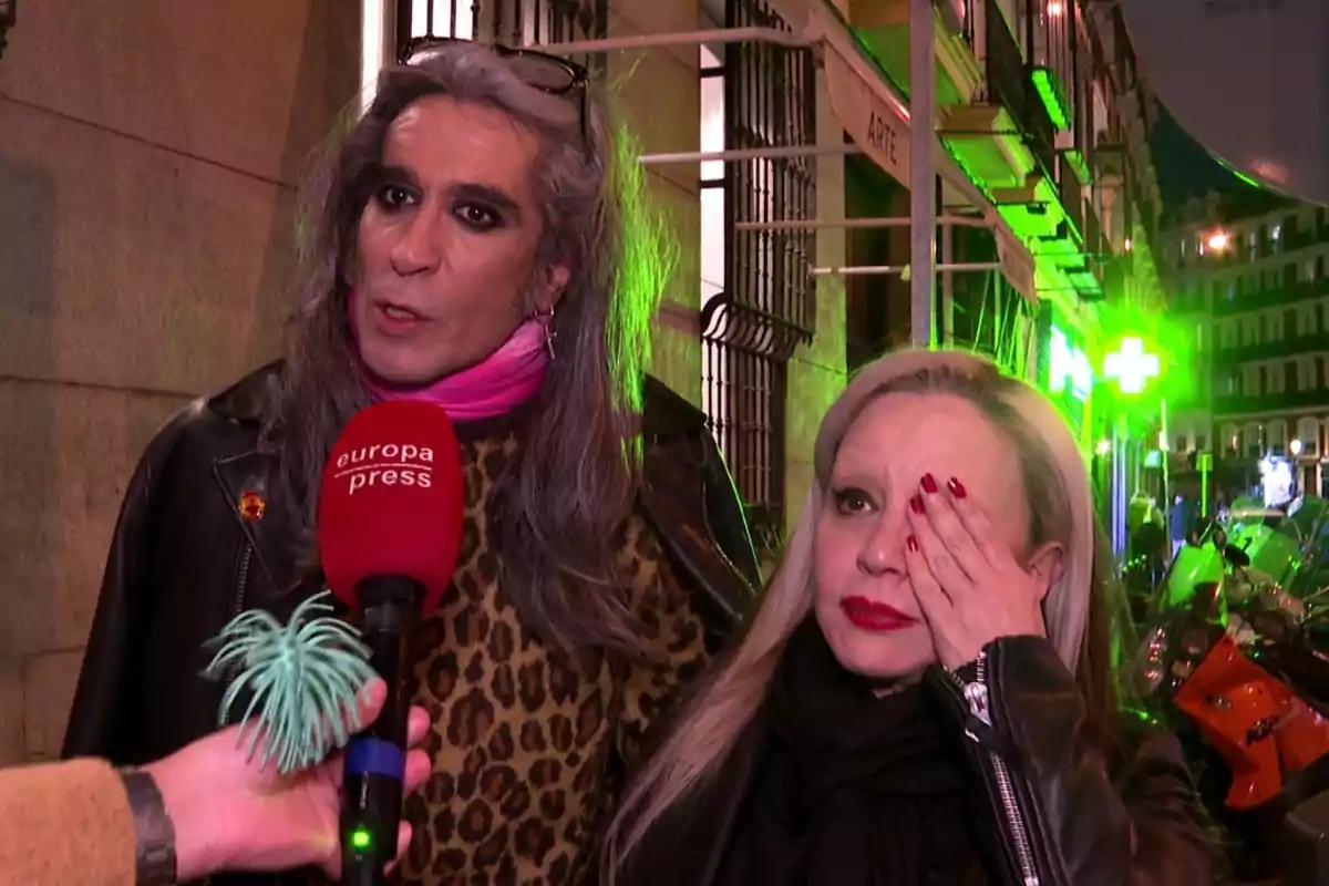 Mario Vaquerizo y Alaska siendo entrevistadas en la calle por un micrófono de Europa Press, una de ellas con el cabello largo y gris y la otra cubriéndose un ojo con la mano, con luces verdes de fondo. Mario Vaquerizo y Alaska siendo entrevistadas en la calle por un micrófono de Europa Press, una de ellas con el cabello largo y gris y la otra cubriéndose un ojo con la mano, con luces verdes de fondo.