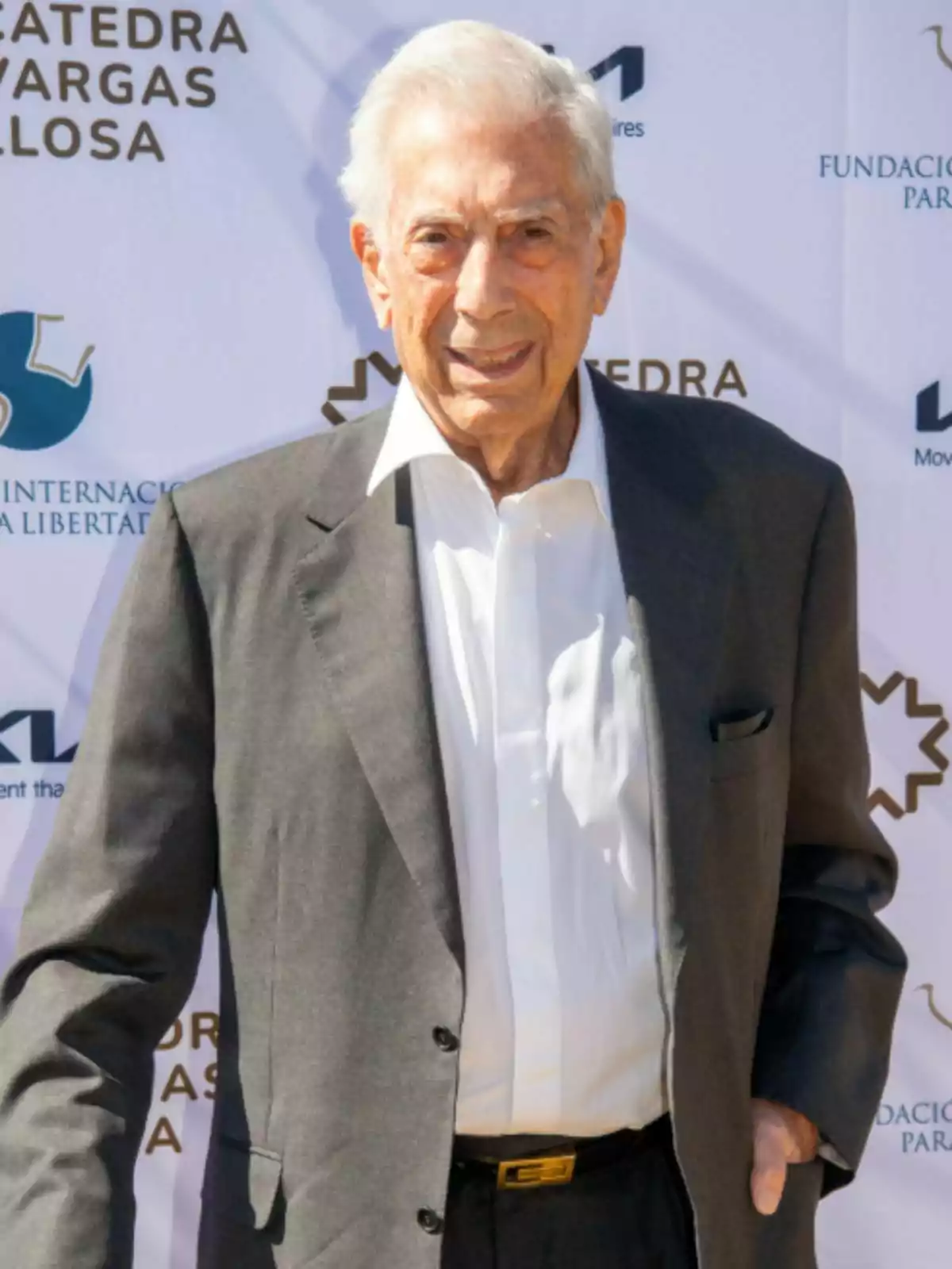 Mario Vargas Llosa con traje oscuro está de pie frente a un fondo con logotipos y texto. Mario Vargas Llosa con traje oscuro está de pie frente a un fondo con logotipos y texto.