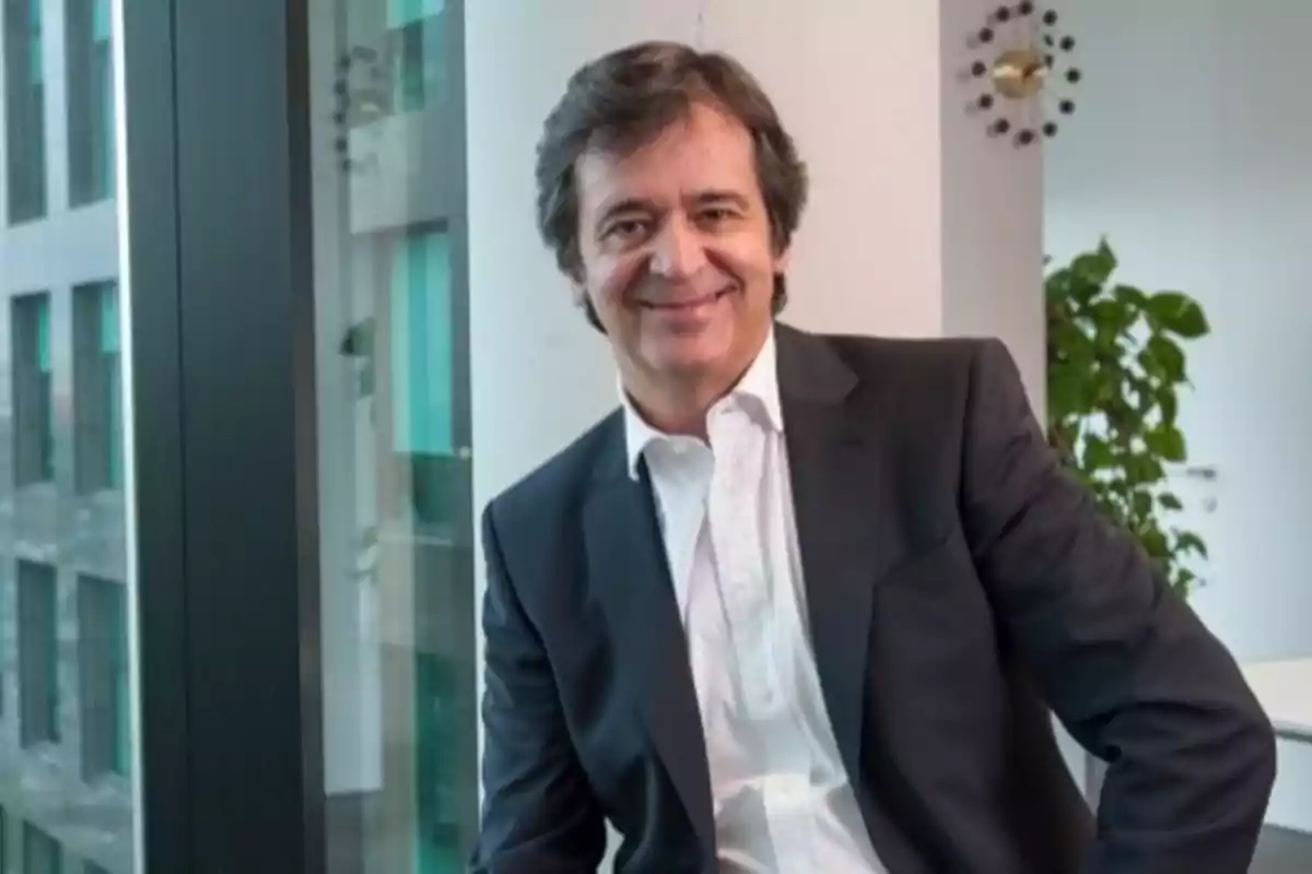 Hombre de mediana edad con cabello oscuro y canoso viste traje oscuro y camisa blanca mientras sonríe sentado junto a una ventana en una oficina moderna