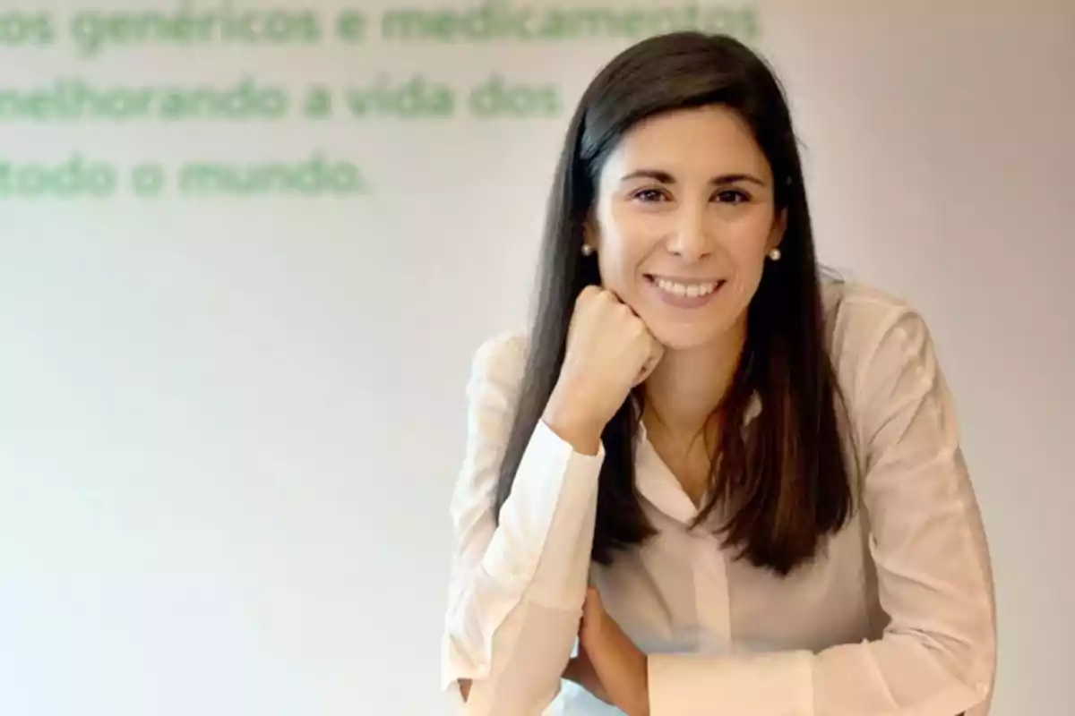 Mujer joven de cabello largo y oscuro, vestida con camisa blanca, sonriendo y apoyando el mentón en su mano, sentada frente a una pared con texto desenfocado al fondo. Mujer joven de cabello largo y oscuro, vestida con camisa blanca, sonriendo y apoyando el mentón en su mano, sentada frente a una pared con texto desenfocado al fondo.