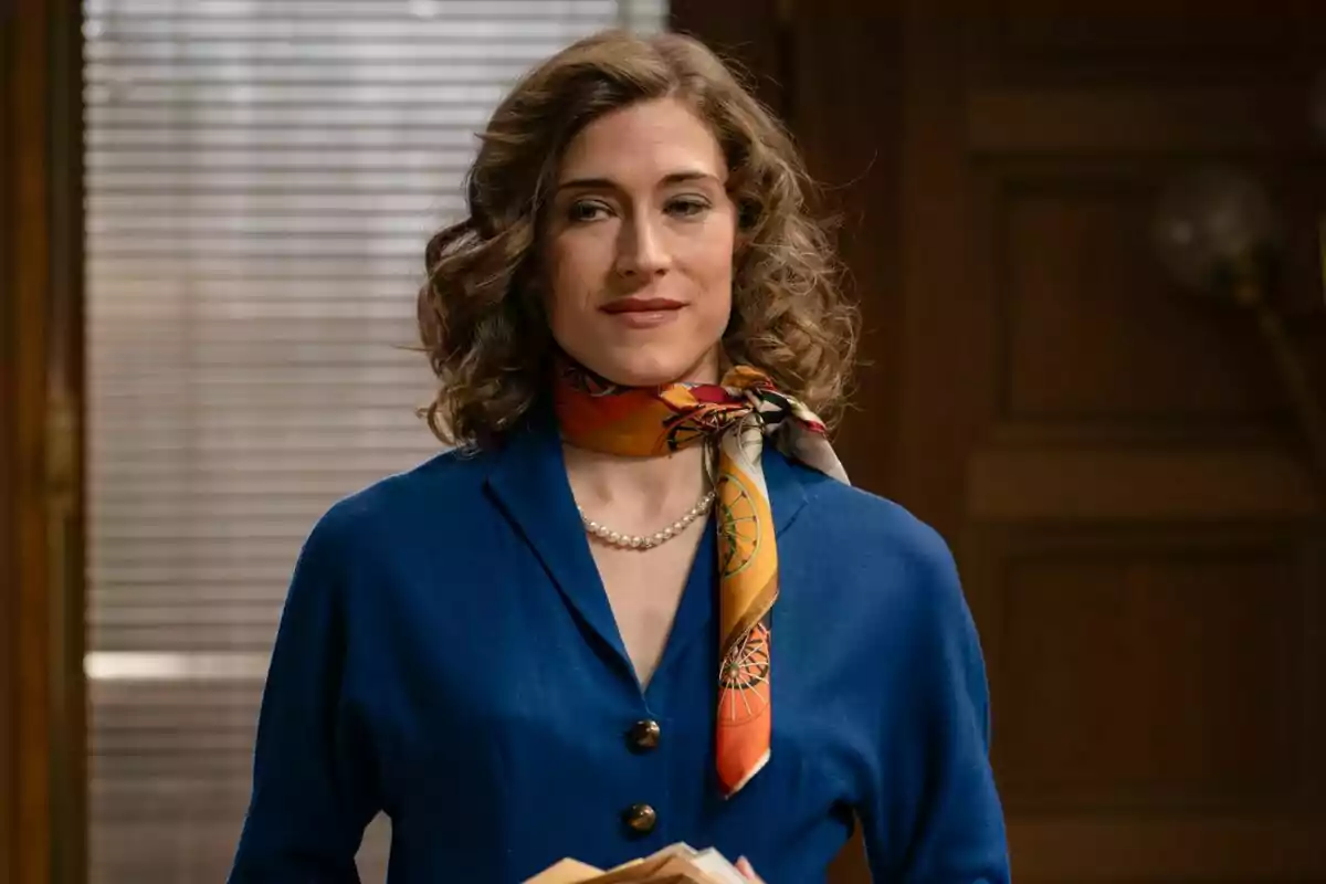 Captura de Marta Belmonte con cabello rizado y castaño, vestida con un traje azul y un pañuelo colorido alrededor del cuello, de pie en una habitación con una ventana de persianas al fondo en Sueños de Libertad Captura de Marta Belmonte con cabello rizado y castaño, vestida con un traje azul y un pañuelo colorido alrededor del cuello, de pie en una habitación con una ventana de persianas al fondo en Sueños de Libertad