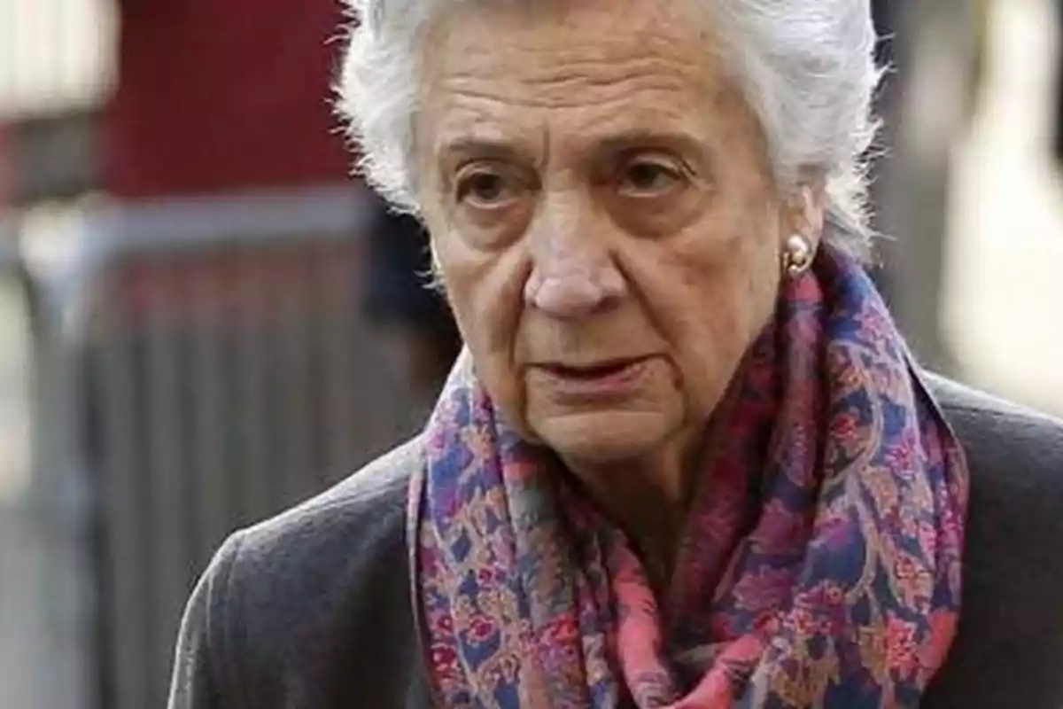 Mujer mayor con cabello canoso y corto, usando una bufanda colorida y una chaqueta oscura.
