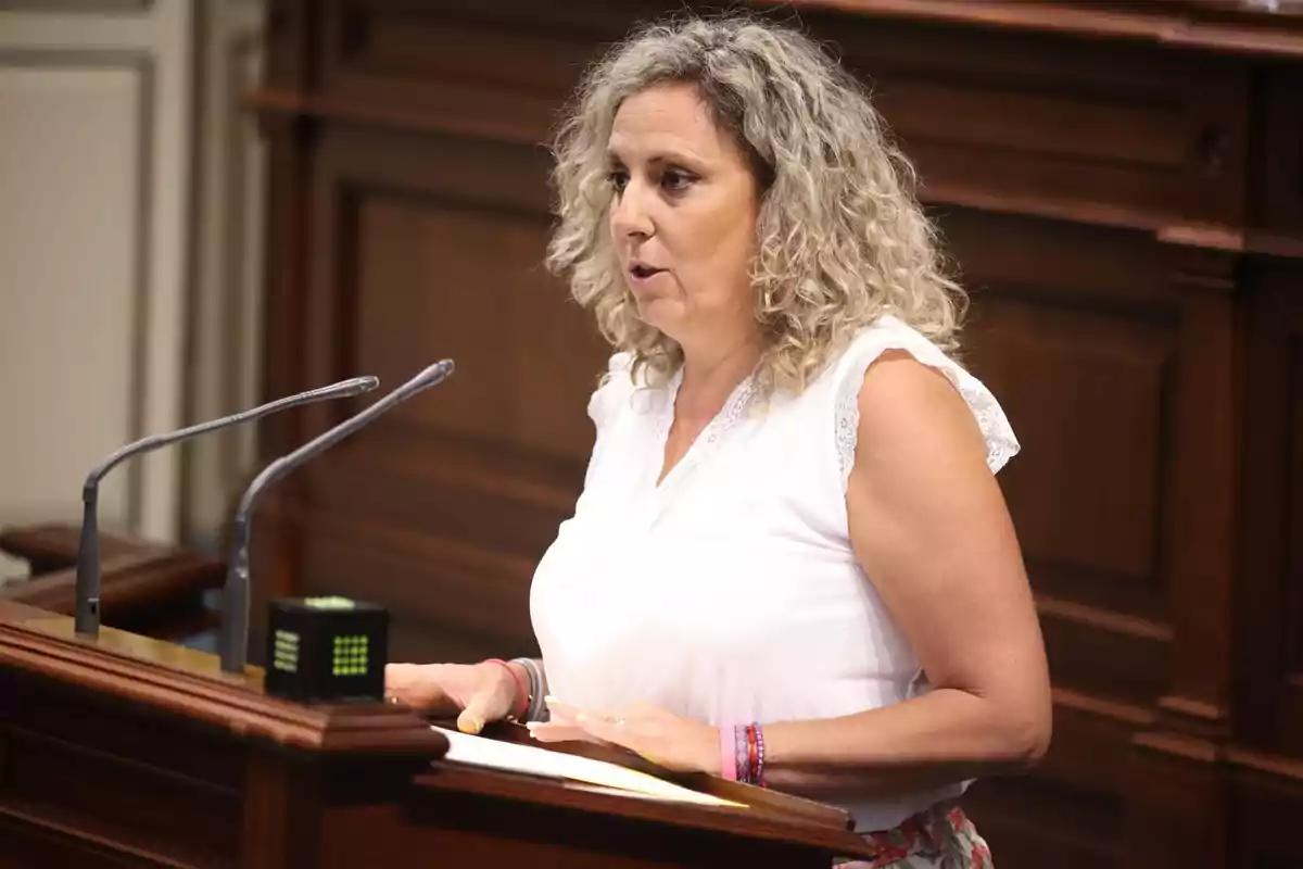 Una mujer de cabello rizado y rubio habla en un podio en un entorno formal.