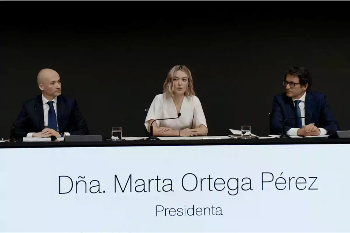 Tres personas en una conferencia, con una mujer en el centro y dos hombres a los lados, con un cartel que dice 