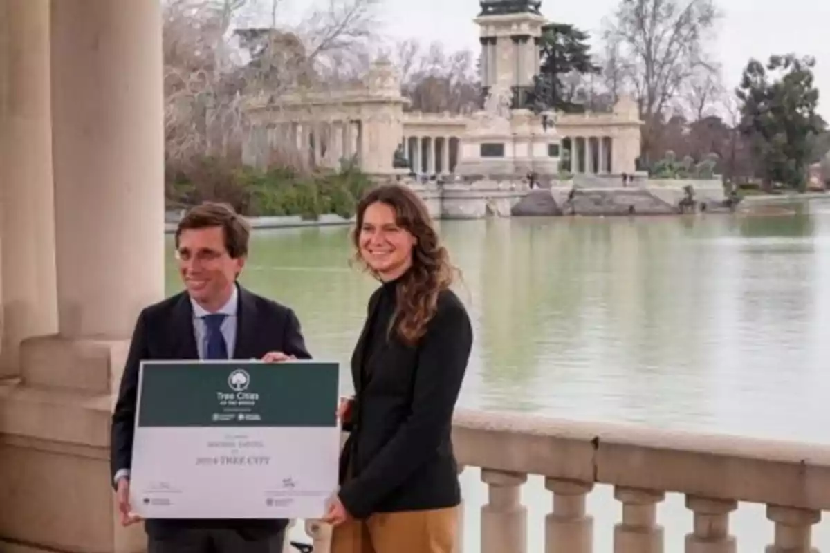 Dos personas sostienen un cartel frente a un lago con un monumento al fondo.