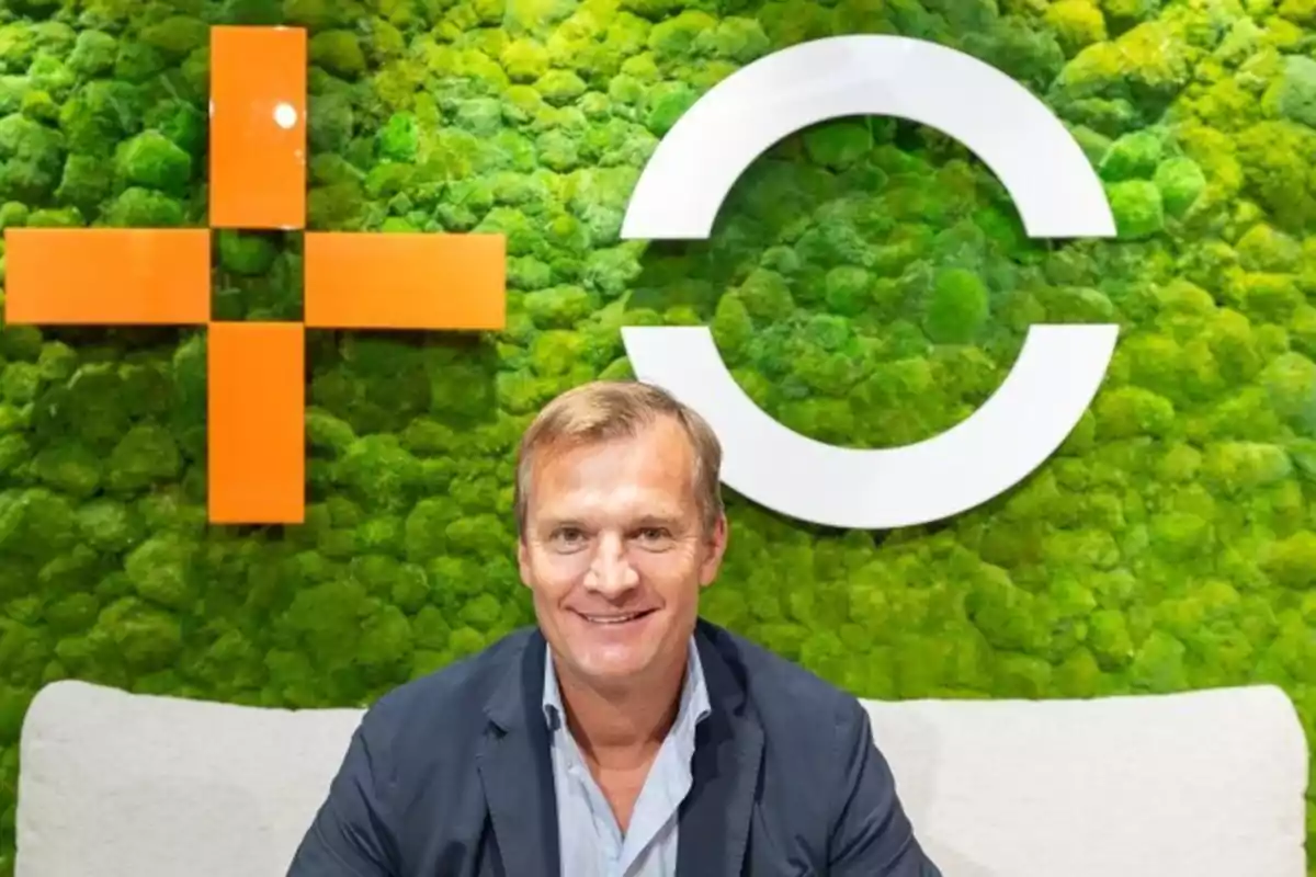 Hombre sonriendo frente a un fondo de musgo verde con símbolos geométricos en naranja y blanco.