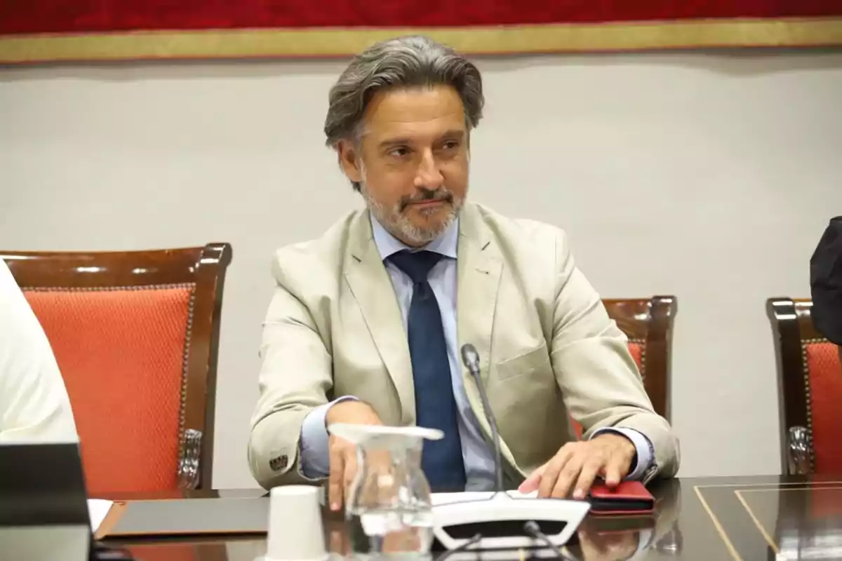 Hombre de cabello canoso y barba sentado en una mesa de reuniones con micrófono y jarra de agua frente a él Hombre de cabello canoso y barba sentado en una mesa de reuniones con micrófono y jarra de agua frente a él