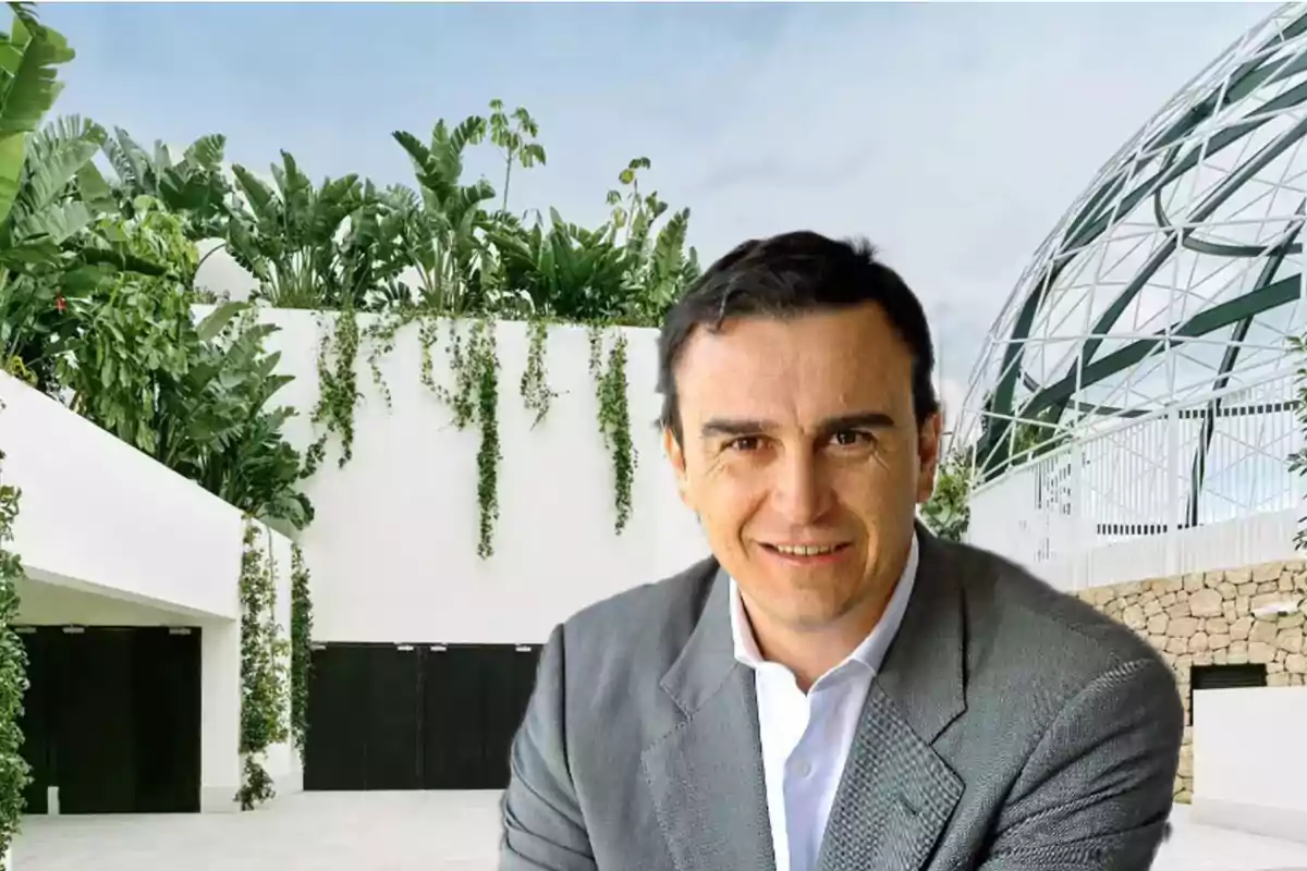 Hombre de cabello oscuro con saco gris sonriendo frente a un edificio moderno con vegetación y una estructura de vidrio.