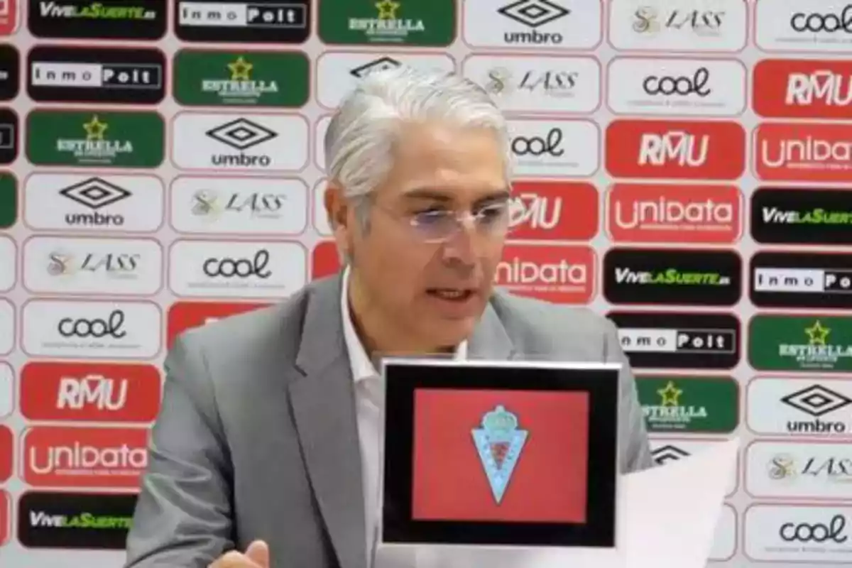 Hombre de cabello canoso y gafas en una conferencia de prensa con un fondo de logotipos de patrocinadores.