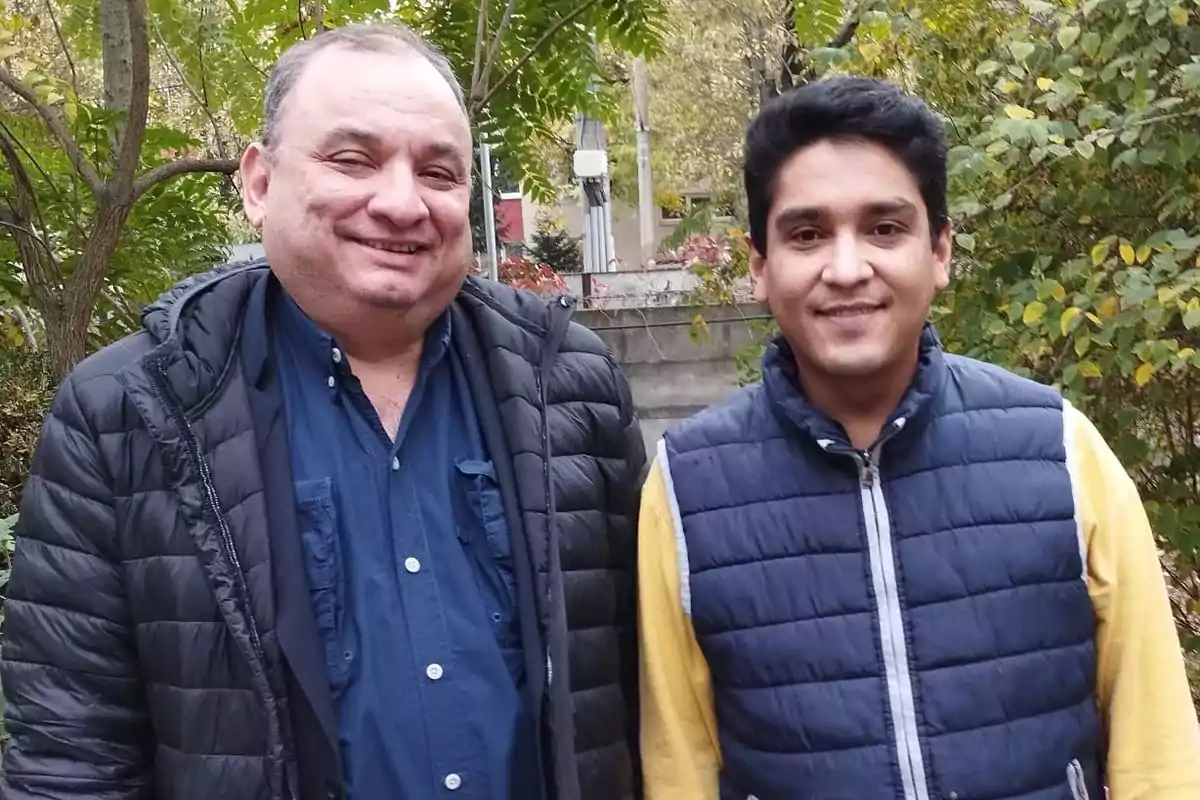 Dos personas posan al aire libre con chaquetas acolchadas y árboles de fondo.