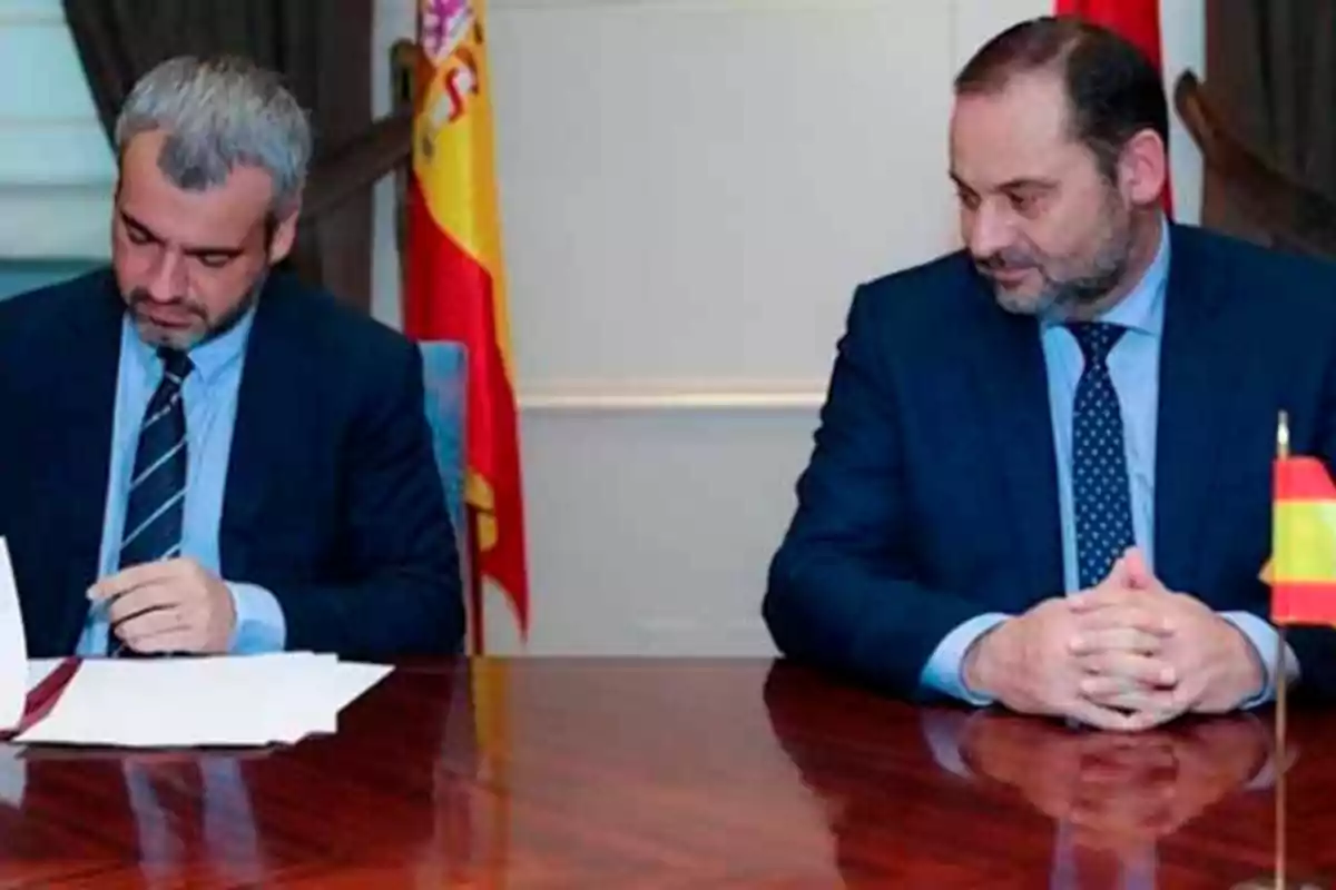 Dos hombres en trajes formales están sentados en una mesa de madera con banderas de España al fondo, uno de ellos está firmando un documento mientras el otro observa.