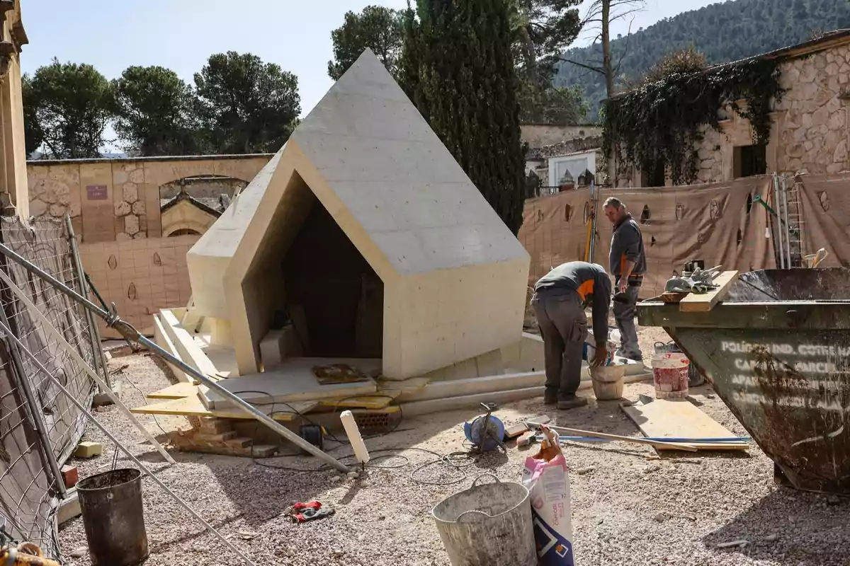 Dos trabajadores en un sitio de construcción junto a una estructura de concreto con forma geométrica en un entorno al aire libre.